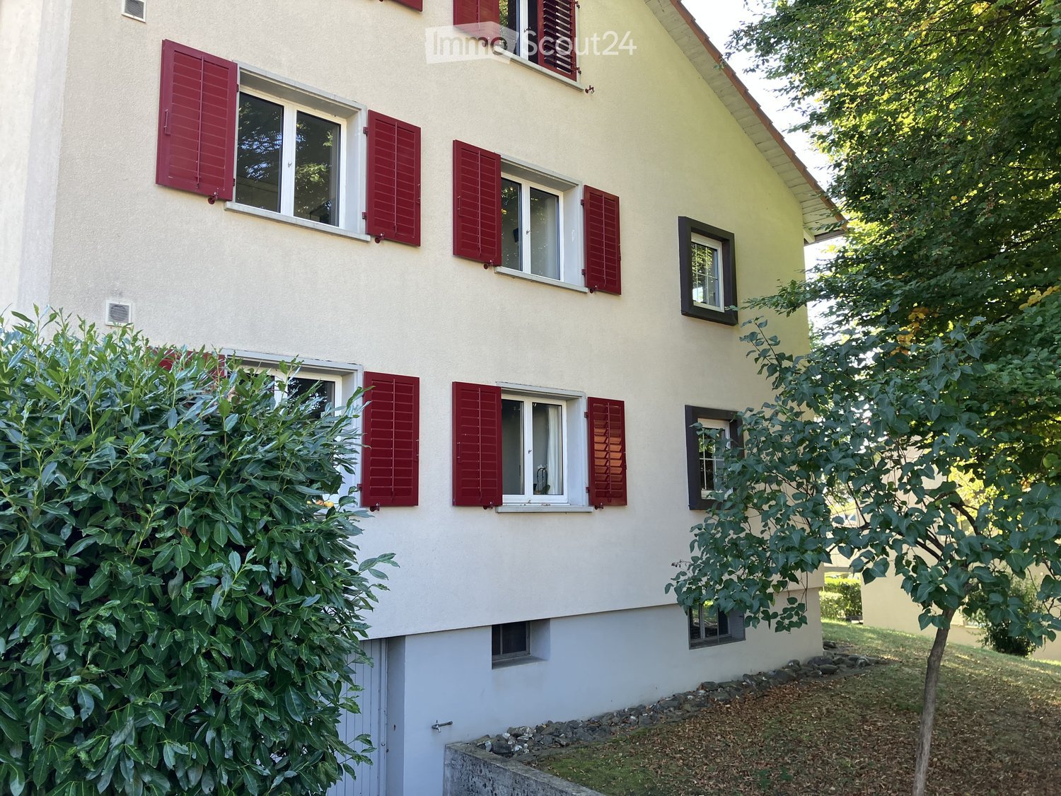 Two-story house with light yellow and white exterior, red shutters, multiple windows, and surrounding greenery.