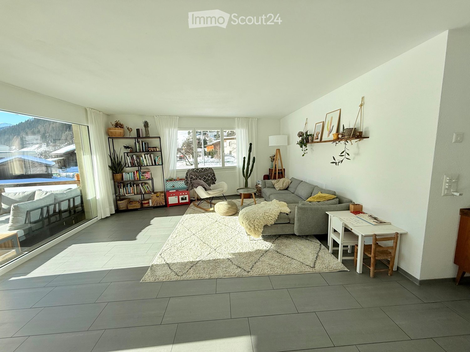 Bright living room with large windows offering a scenic view, featuring a grey sectional sofa, a white armchair, a small round table, a bookshelf, a floor lamp, and a white shaggy rug.