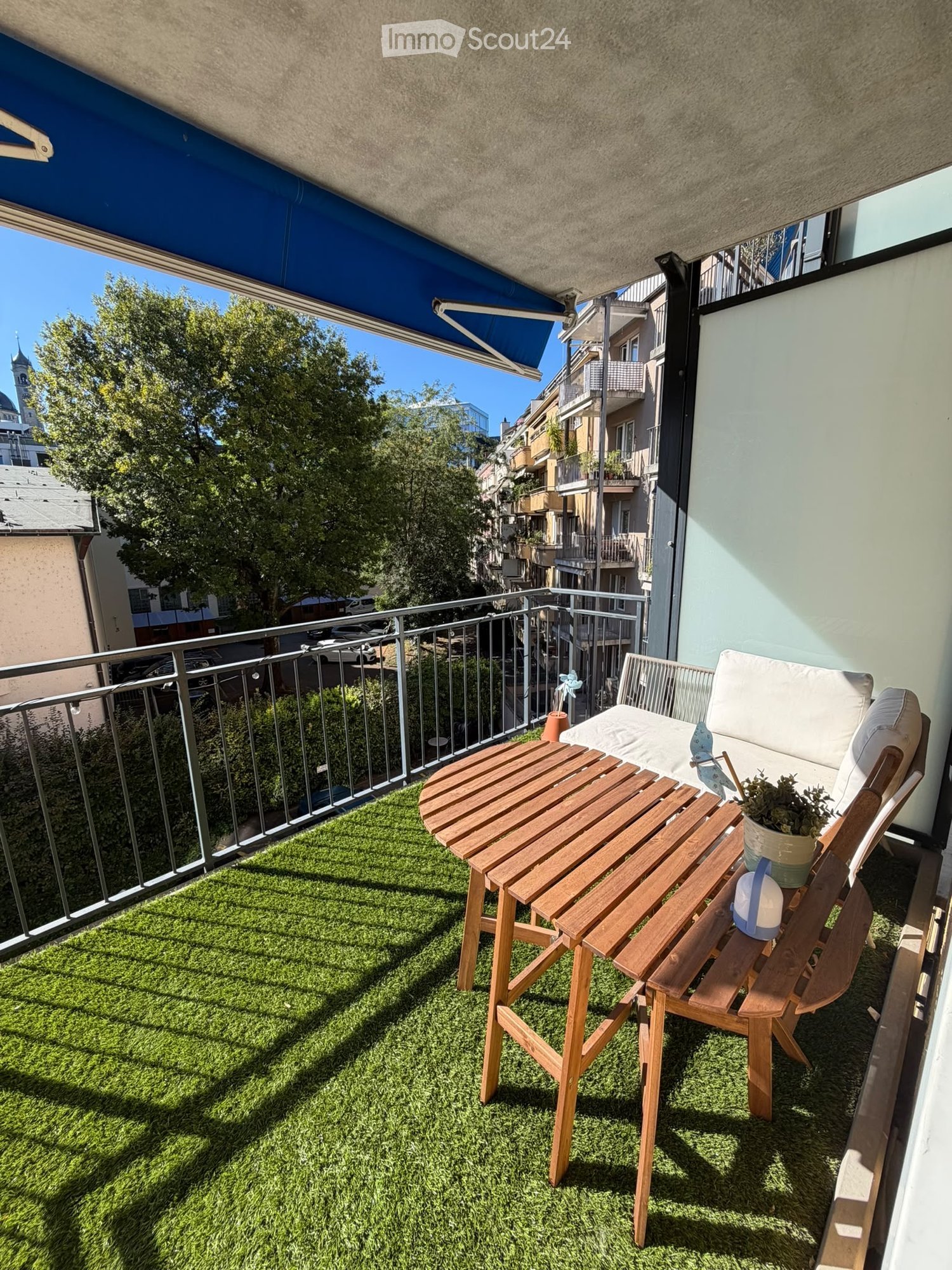 Balcony with artificial grass, wooden table, chair, and potted plant, overlooking a cityscape with buildings and trees.