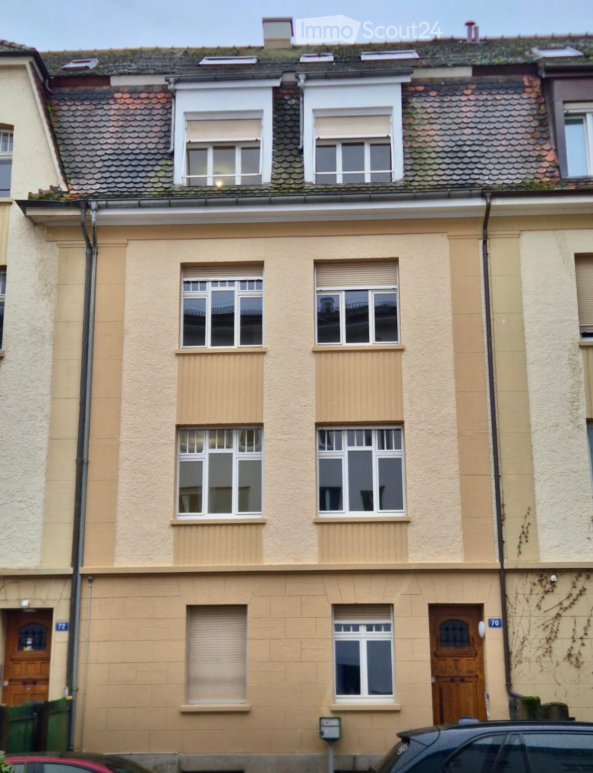 Two-story residential building with beige facade, multiple windows with shutters, and wooden doors.
