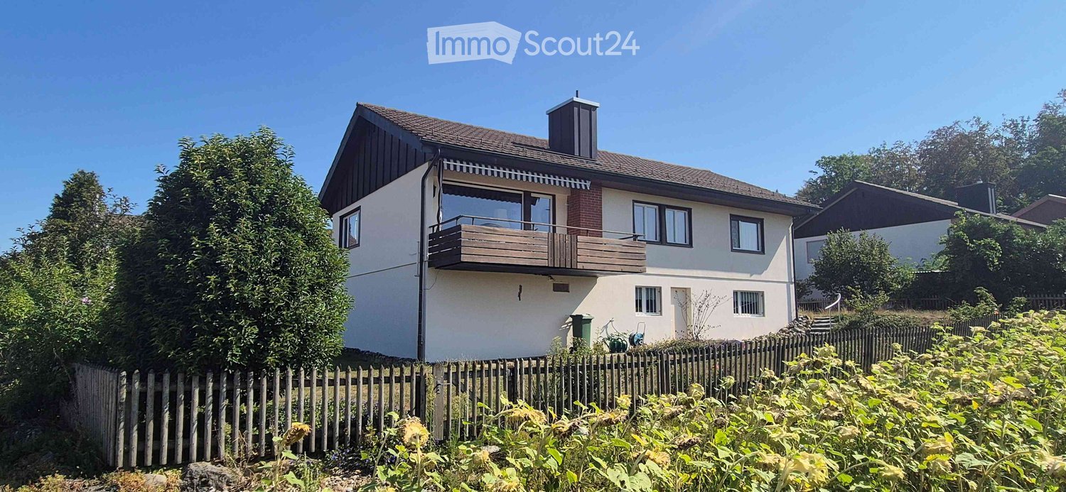 Two-story house with a balcony, surrounded by greenery and a wooden fence.