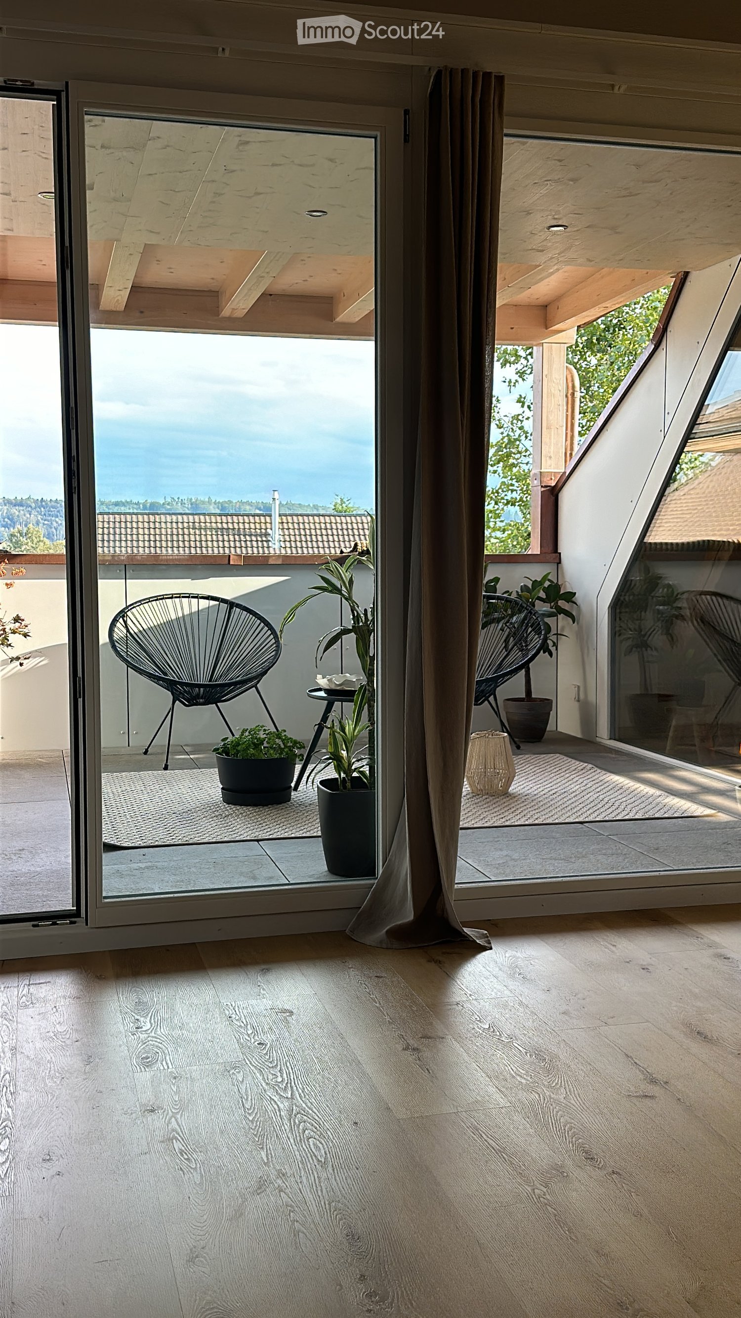 Modern balcony with wooden floor, large glass doors, two black chairs, small table, and potted plants.