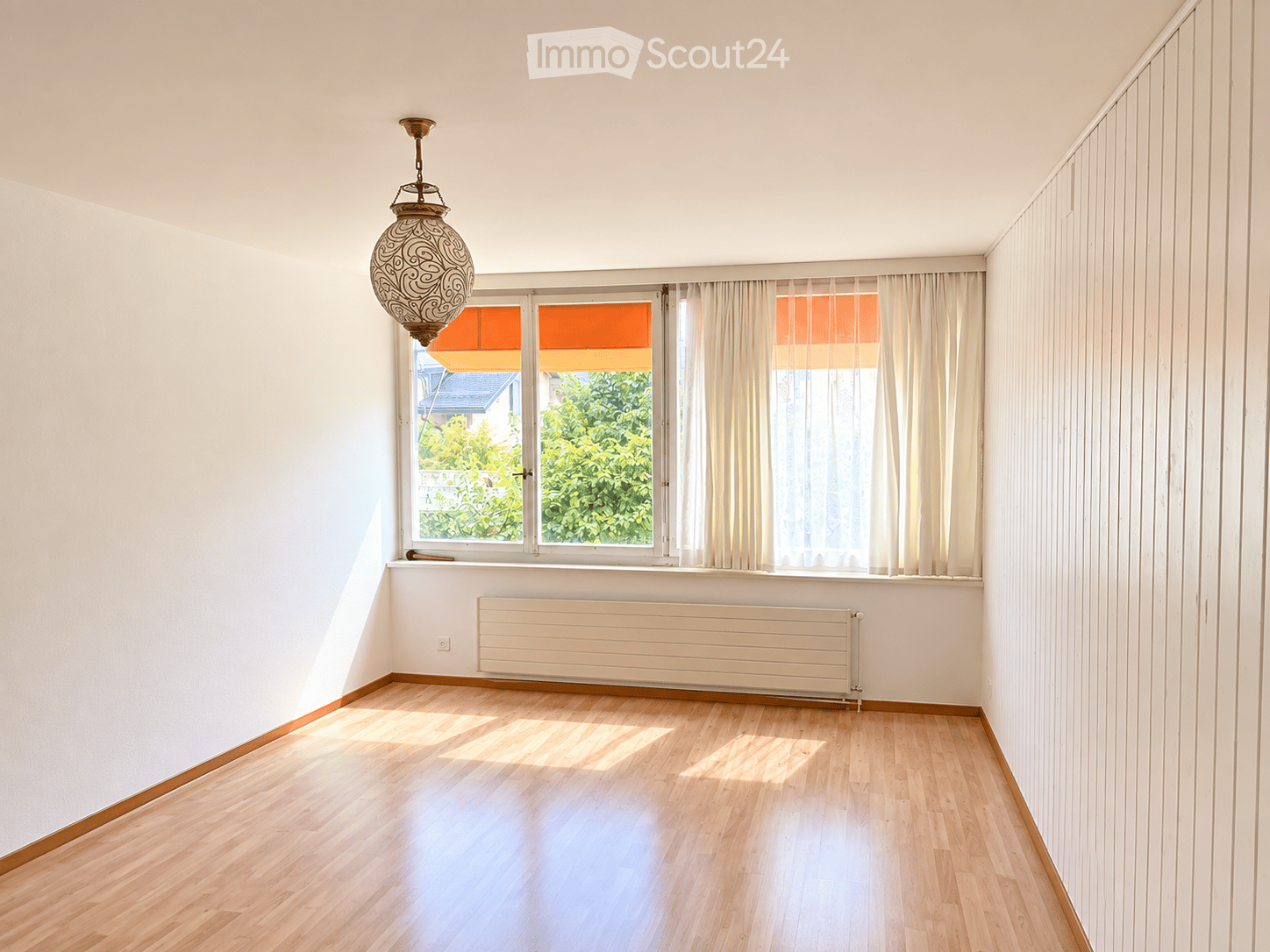 Empty room with light wooden flooring, white walls, large windows with white curtains, and a decorative hanging light fixture.