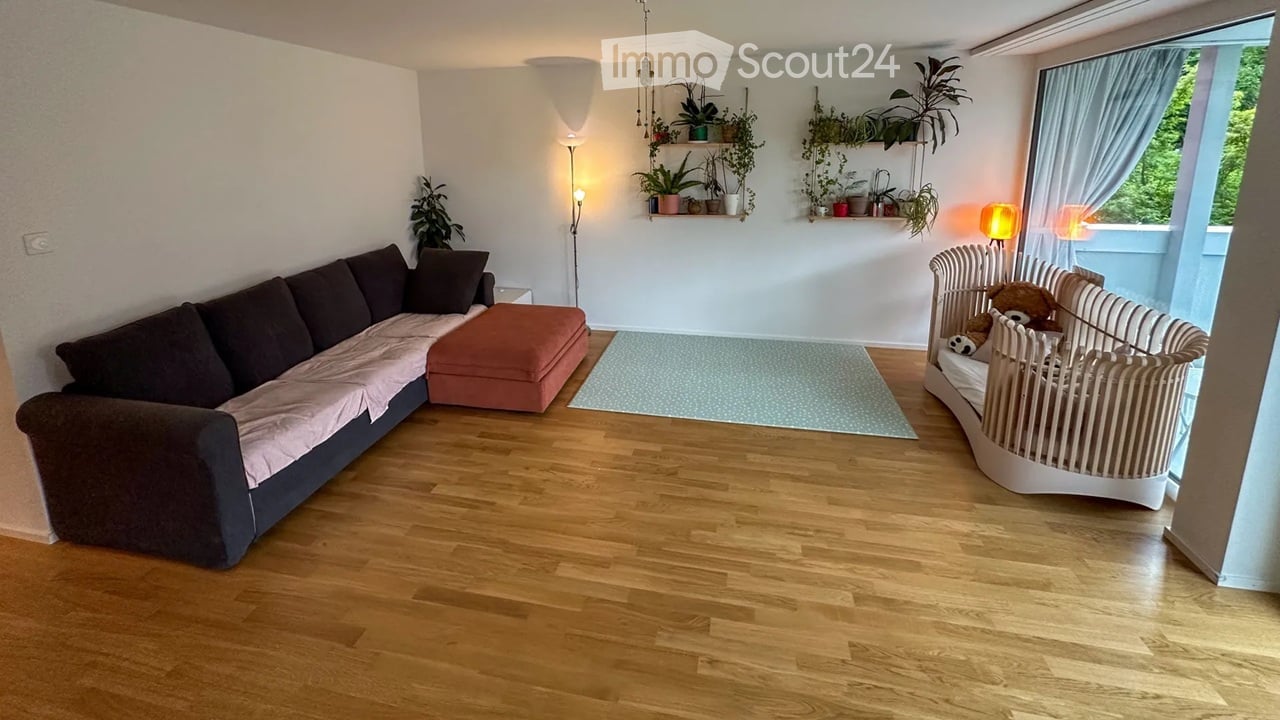 Living room with wooden flooring, a dark brown sectional sofa with pink throw, an orange ottoman, a white baby crib with stuffed animals, floor lamp, table lamp, and wall-mounted shelves with plants.