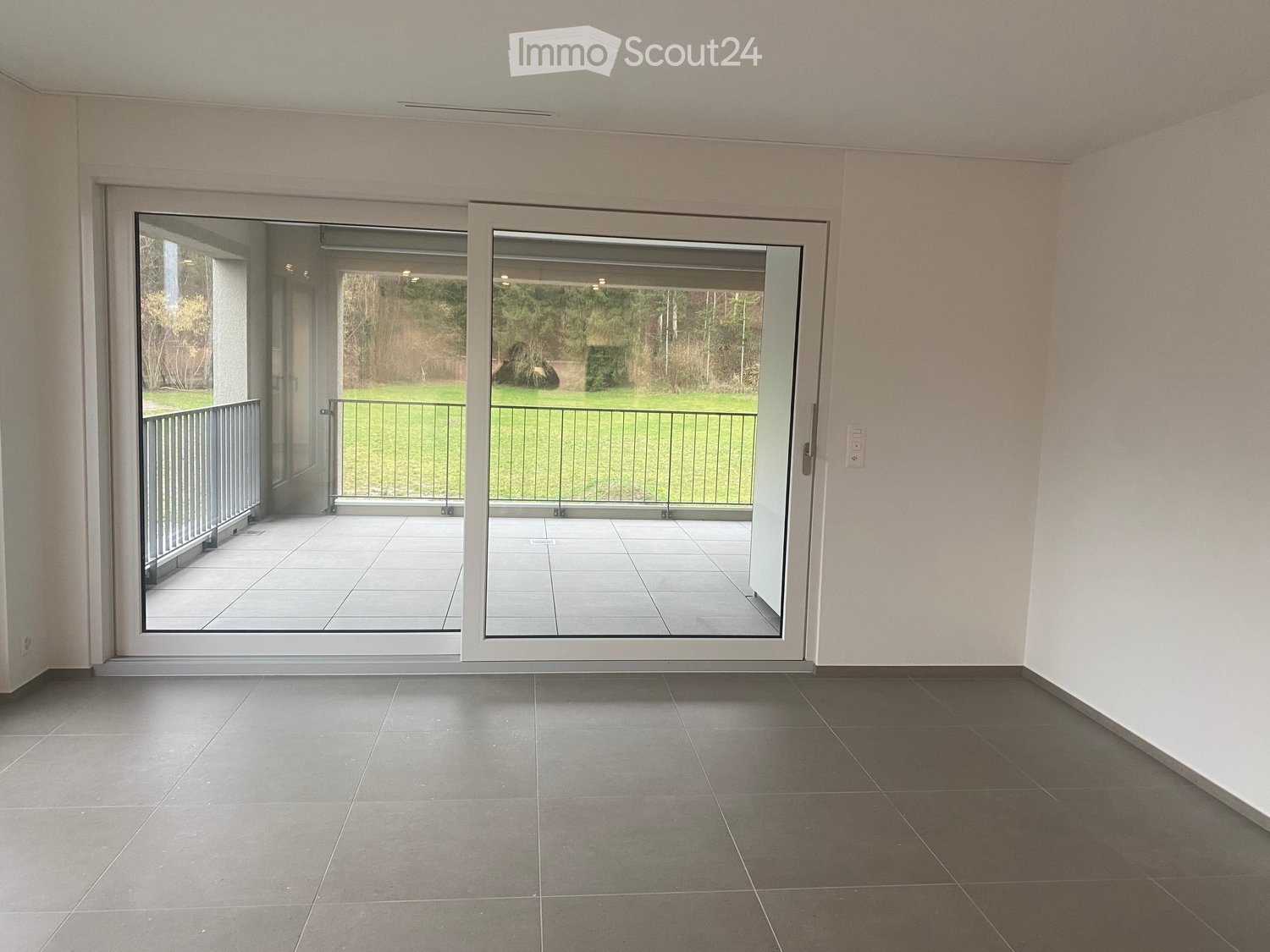 Empty room with grey tiled flooring, white walls, and large sliding glass doors leading to a balcony with a metal railing and a view of a green outdoor area.
