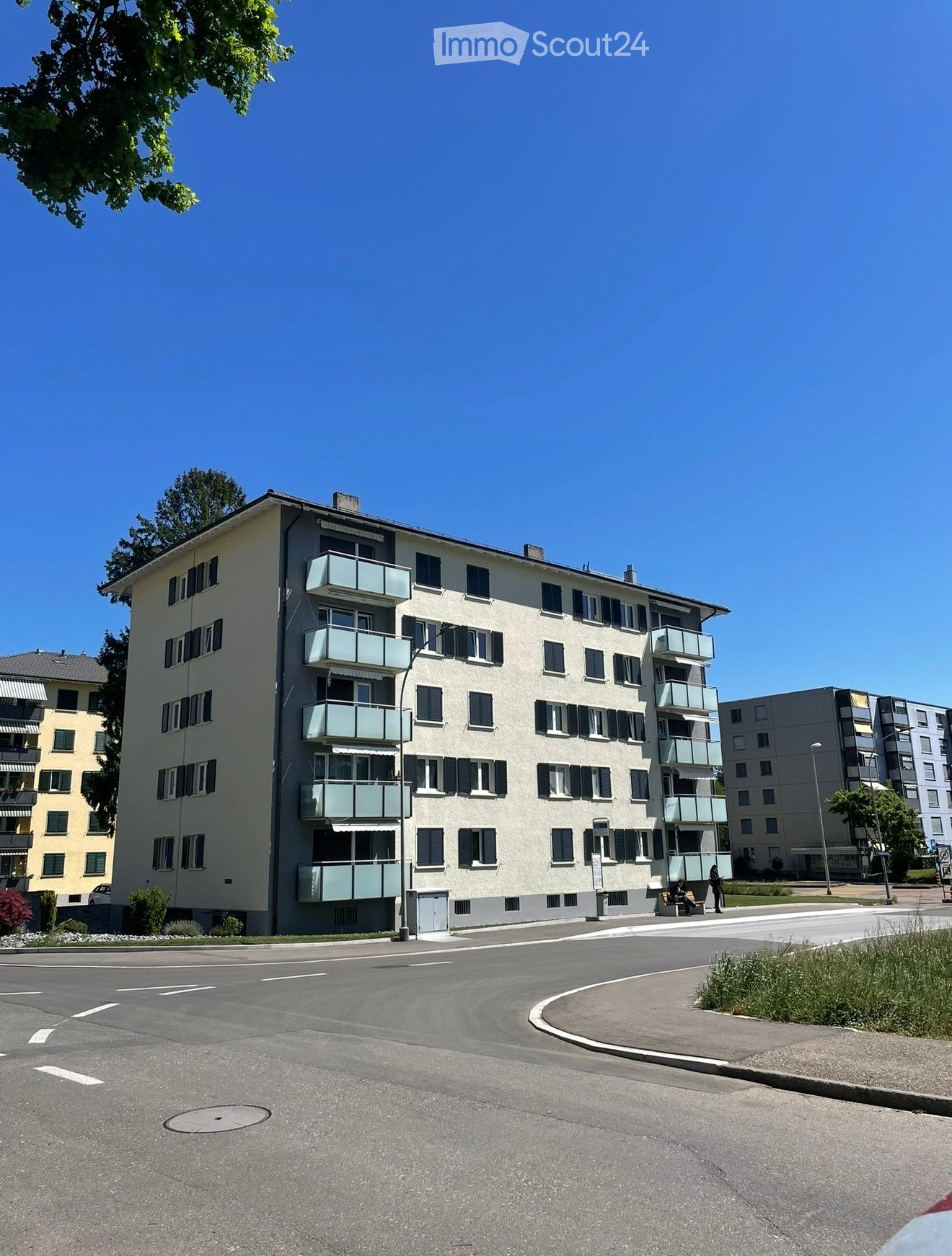 Multi-story residential building with balconies, windows, and shutters, surrounded by a paved area and greenery.