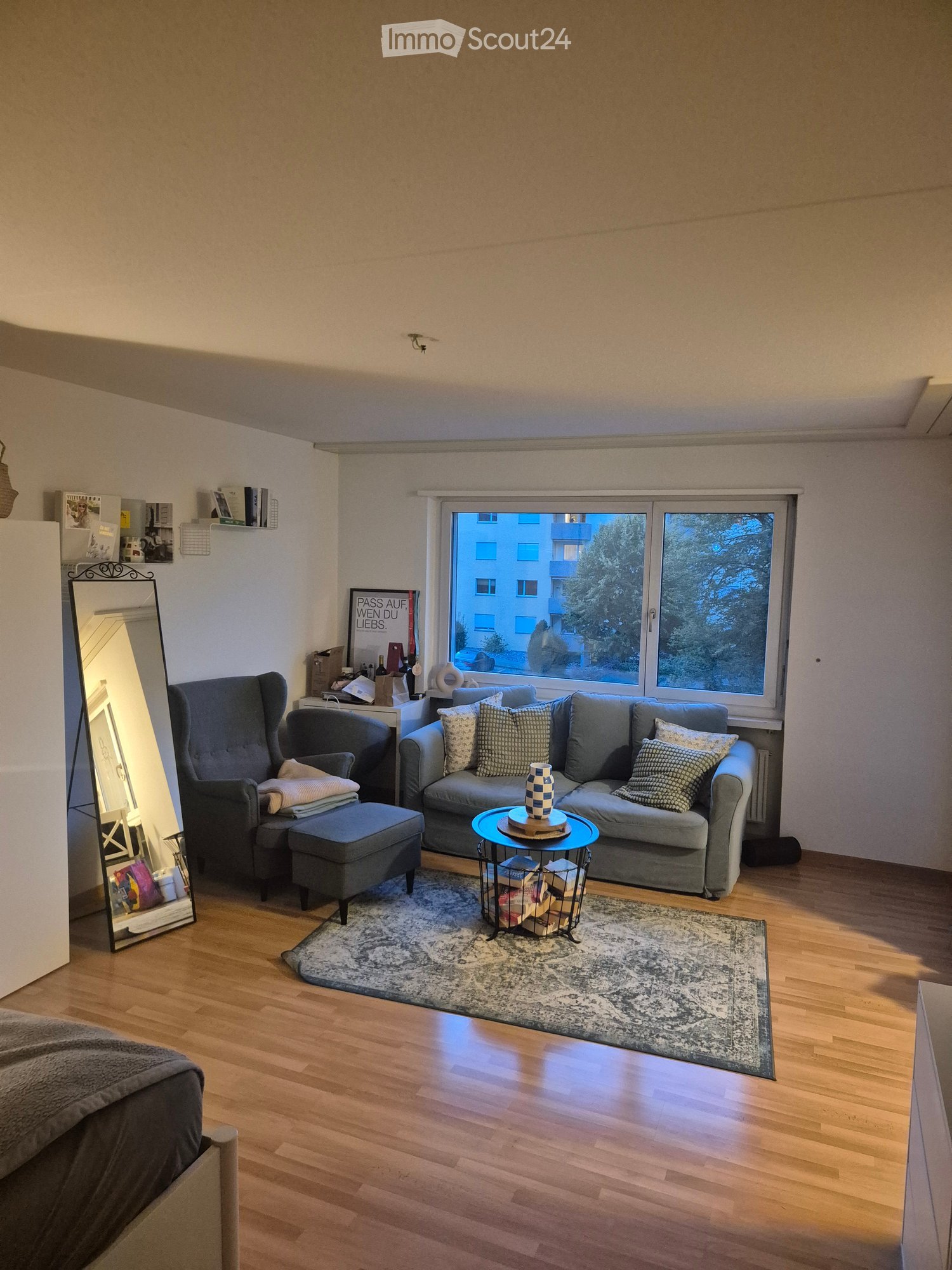 Modern living room with wooden flooring, large window, grey sofa, coffee table, ottoman, armchair, and a decorative mirror.