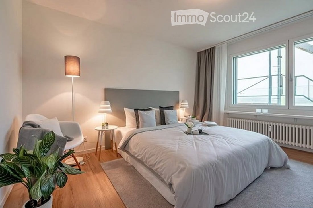 Modern bedroom with a large bed, nightstands, a chair, a small table, and a floor lamp. The room features large windows with curtains and a potted plant near the chair.