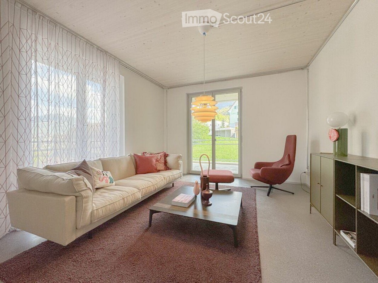 Modern living room with a white sofa, coffee table, pink armchair, and a hanging lamp. The room features large windows with sheer curtains and a view of greenery outside.