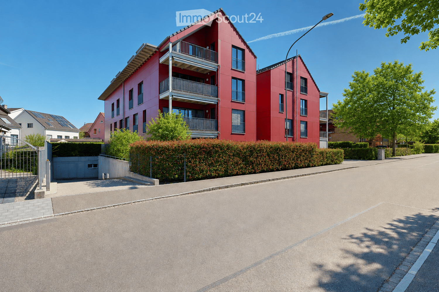 A modern red apartment building with balconies, surrounded by greenery and a paved driveway.