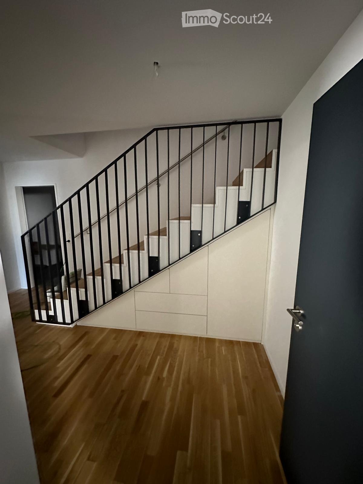 Modern hallway with wooden flooring, white walls, and a staircase with a black metal railing leading to an upper level.