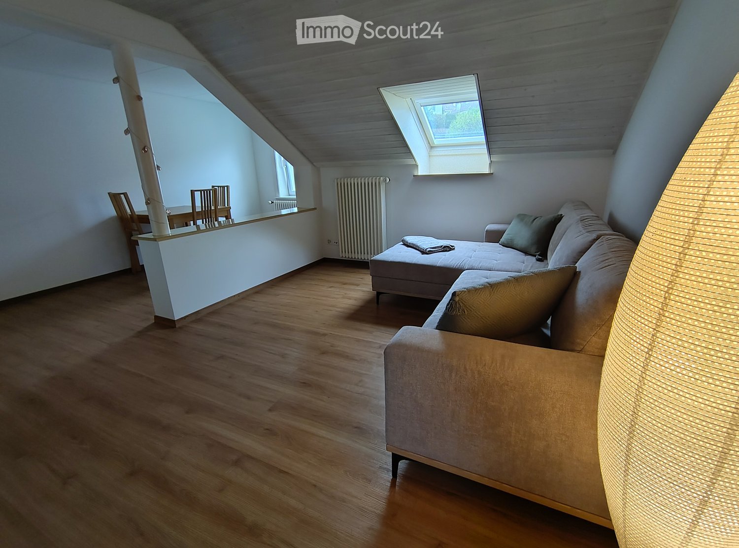 Living room with wooden flooring, a grey sectional sofa, a small dining area with a table and chairs, and a floor lamp with a textured shade.