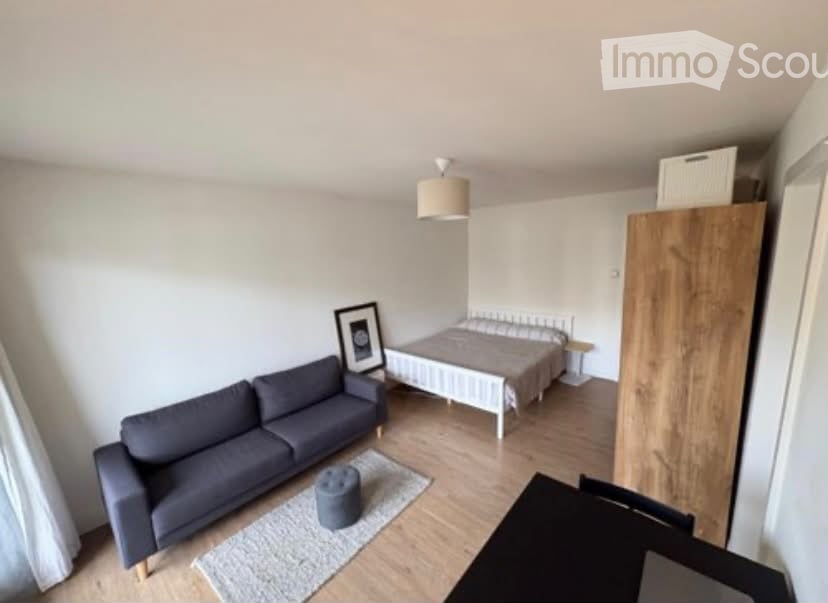 A modern, compact living space with a sofa, a small bed, a wardrobe, a picture frame, and a small round stool. The room features light wooden flooring and white walls.