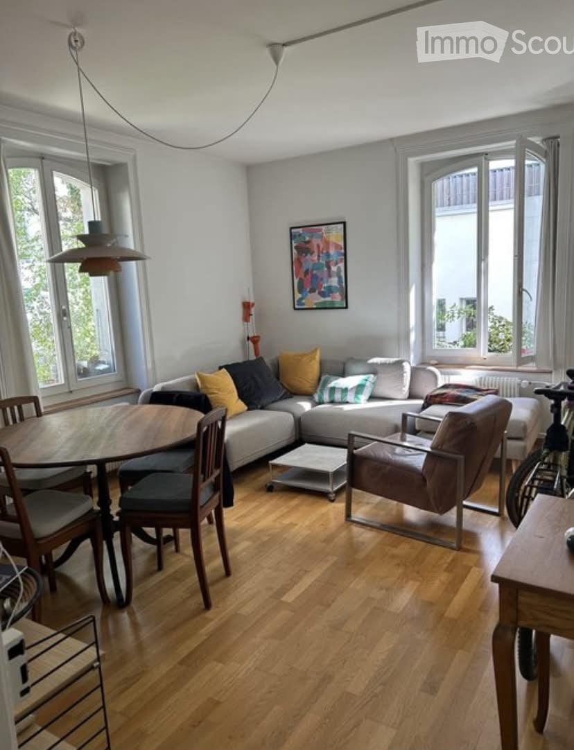 Bright living room with light grey sectional sofa, colorful throw pillows, wooden dining table with chairs, pendant light, and artwork on the wall.