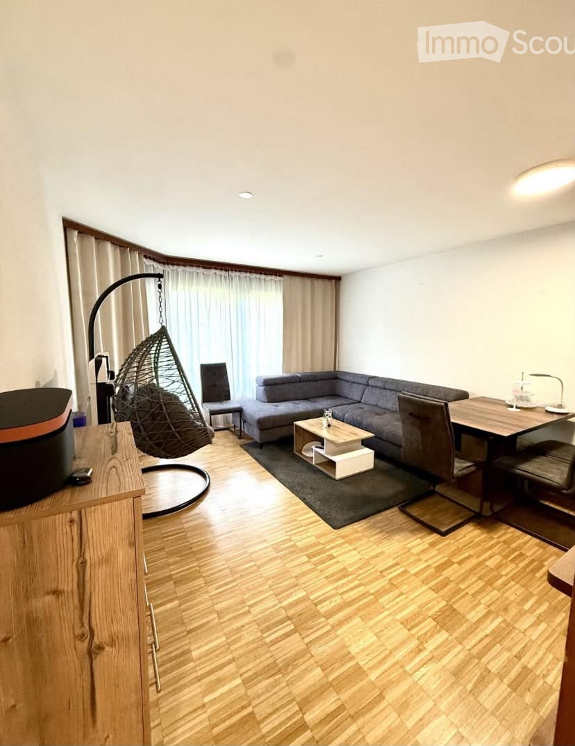 A modern living room with wooden flooring, a grey sectional sofa, a coffee table, a wicker hanging chair, a dining table with chairs, and a wooden cabinet.