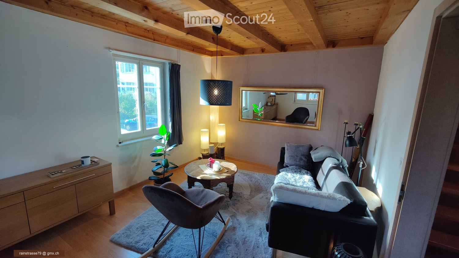 Modern living room with exposed wooden ceiling beams, large window, grey sofa, coffee table, armchair, sideboard, and decorative items.