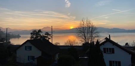 View of a serene lakeside scene with houses, mountains, and a sunset over the water.