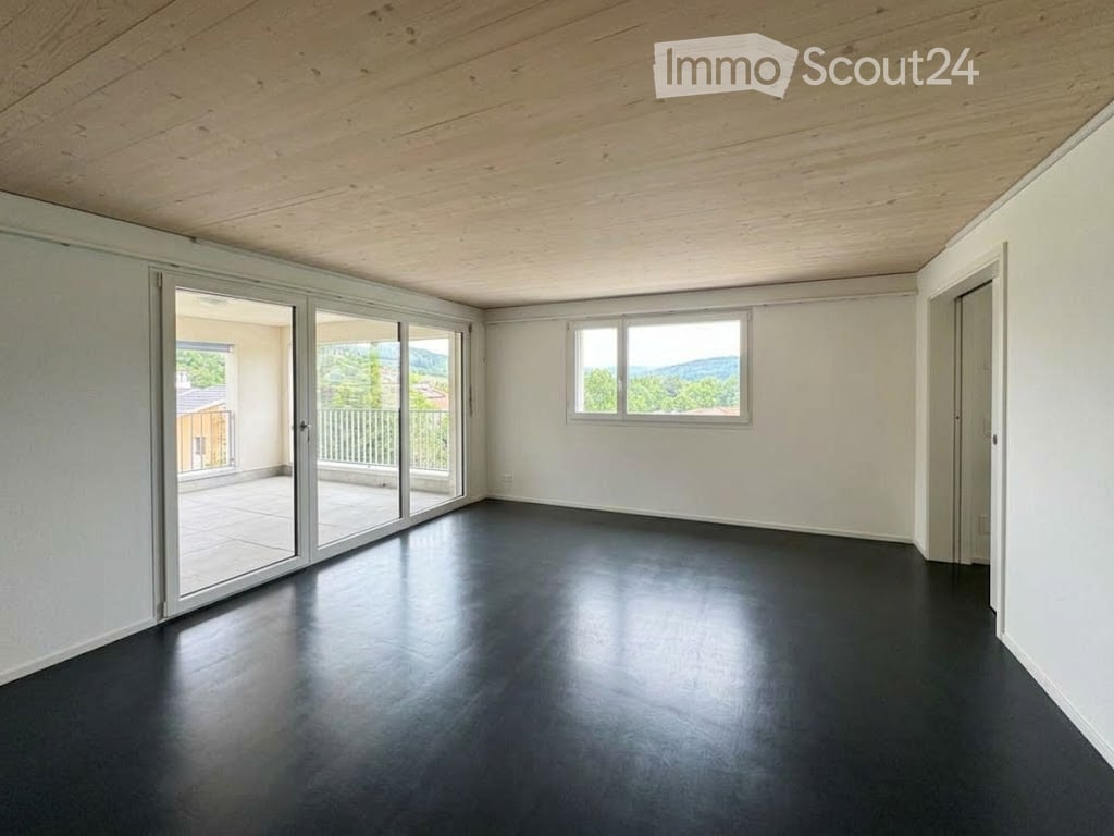 Empty room with white walls, dark flooring, large sliding glass doors leading to a balcony with a scenic view, and a window with a view of greenery and mountains.