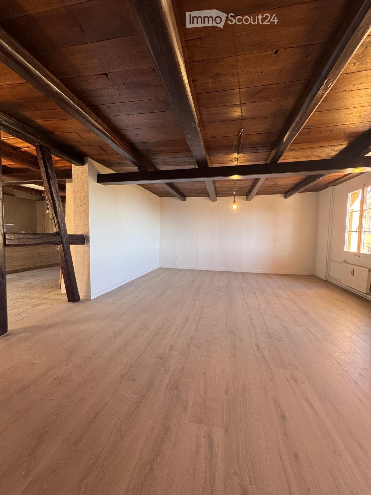 Empty room with wooden flooring, exposed wooden beams on the ceiling, white walls, and a single window letting in natural light.