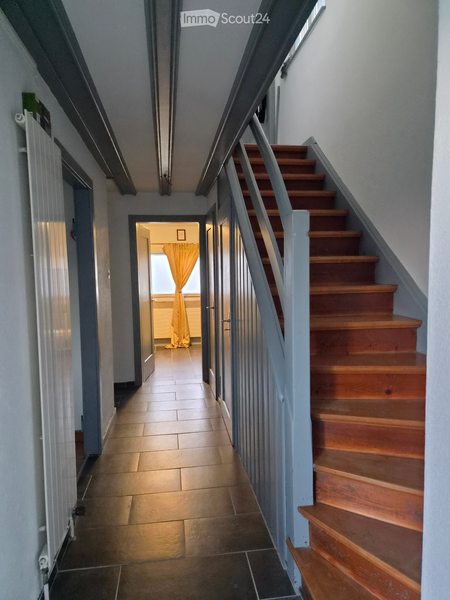 A narrow hallway with tiled flooring, a wooden staircase with a metal railing, and an open doorway leading to another room with a yellow curtain.