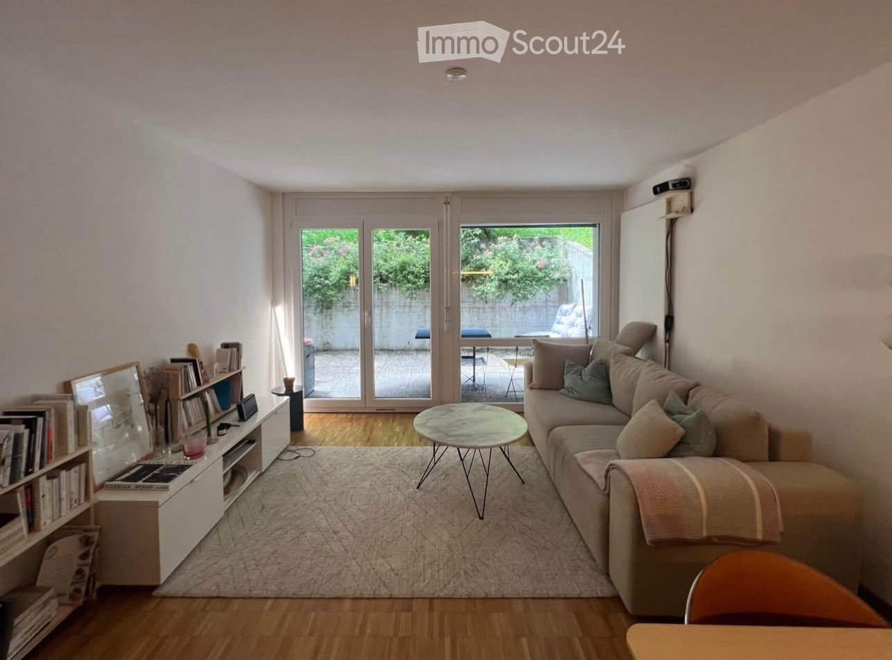 Bright living room with wooden flooring, large sliding glass doors leading to a balcony, a beige sofa with multiple cushions, a round coffee table, a bookshelf, and a small table with a chair.