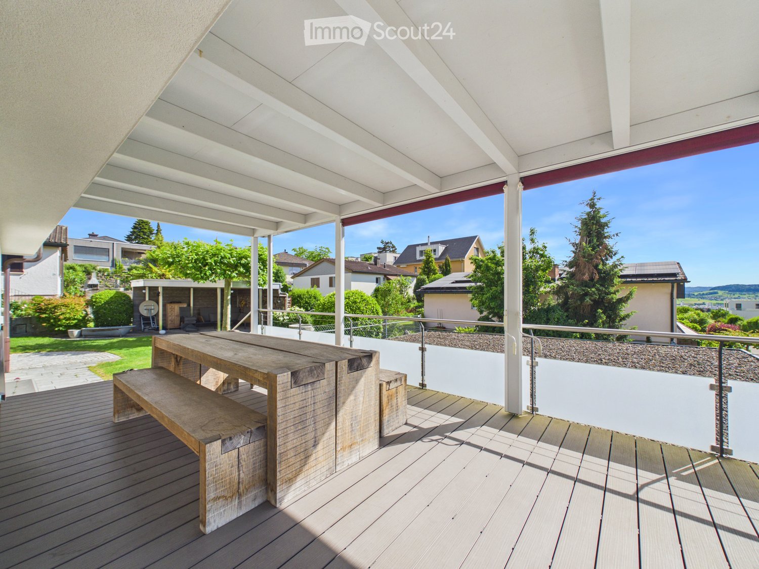 Wooden deck with a large table and bench, metal railing, and a view of a suburban neighborhood with houses and trees.