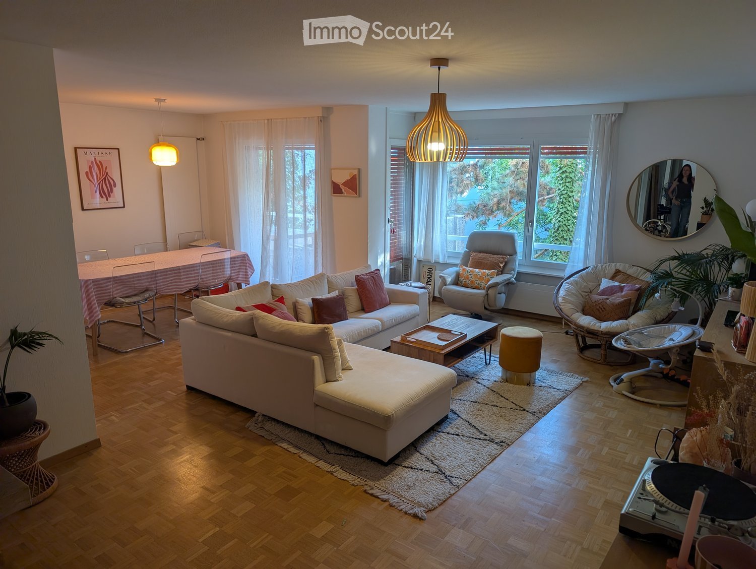 Bright living room with wooden flooring, large windows, a white sectional sofa, a coffee table, a dining area with a table and chairs, and decorative plants.