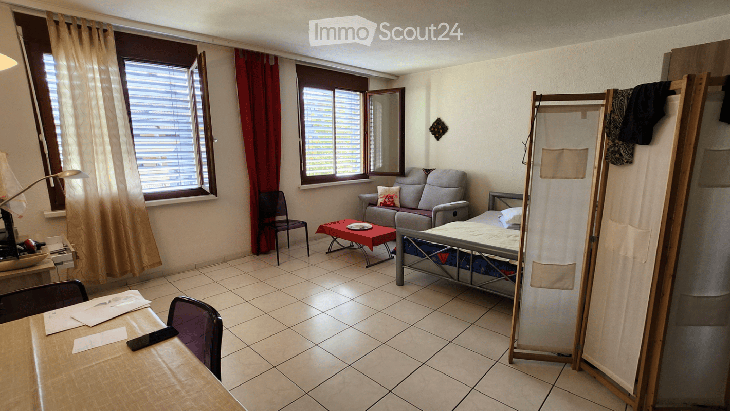 A small, furnished room with a sofa, a bed, a table, and a chair. The room has tiled flooring, large windows with blinds, and a mirror on the wall.