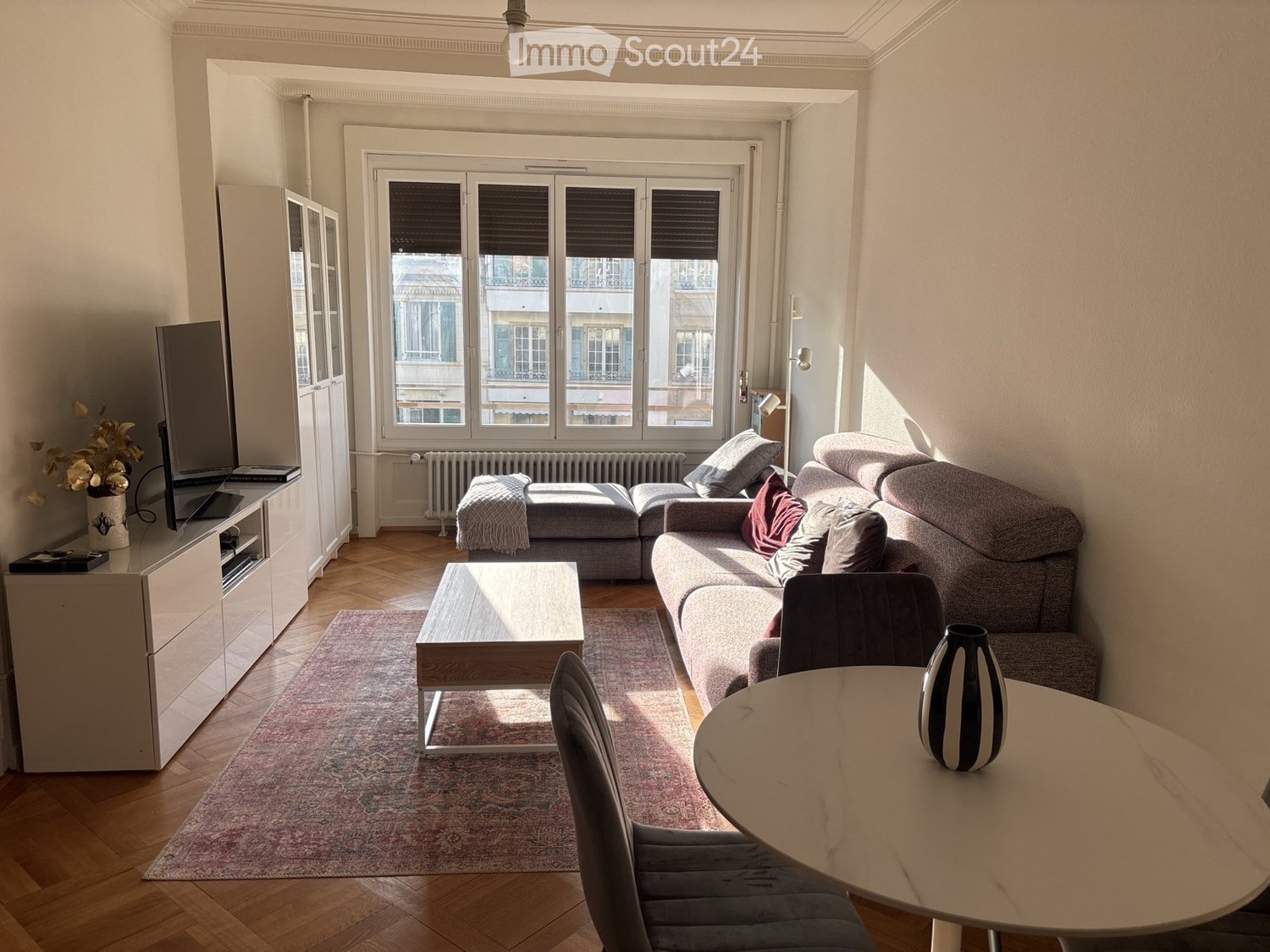 Bright living room with wooden flooring, large windows, grey sectional sofa, coffee table, dining table with chairs, and a TV cabinet.