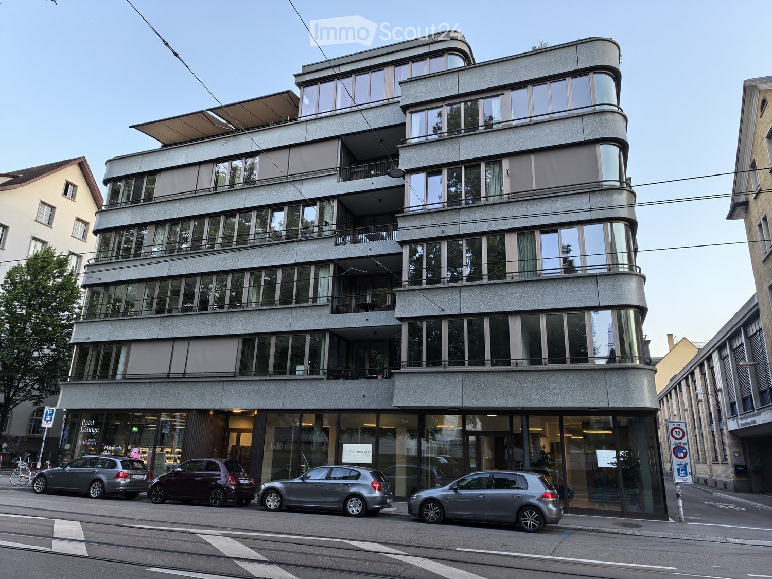 Modern multi-story building with large windows and balconies, located on a street with parked cars and a bike lane.