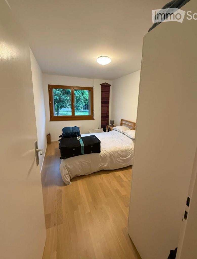 Cozy bedroom with light wooden flooring, a large window with wooden frame, a single bed with white bedding, a nightstand with a lamp, and luggage placed on the floor.