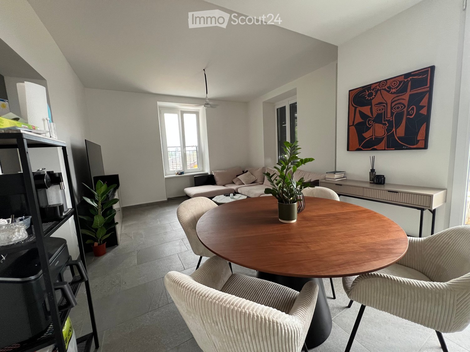 Modern living room with a round wooden table, beige chairs, sofa, potted plants, and a TV screen on a stand.