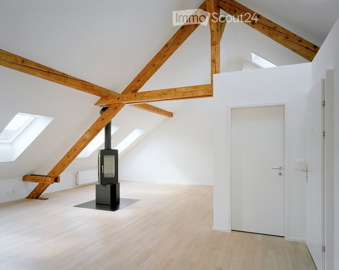 Bright, empty attic room with wooden beams, light wooden flooring, white walls, skylights, and a black wood-burning stove.