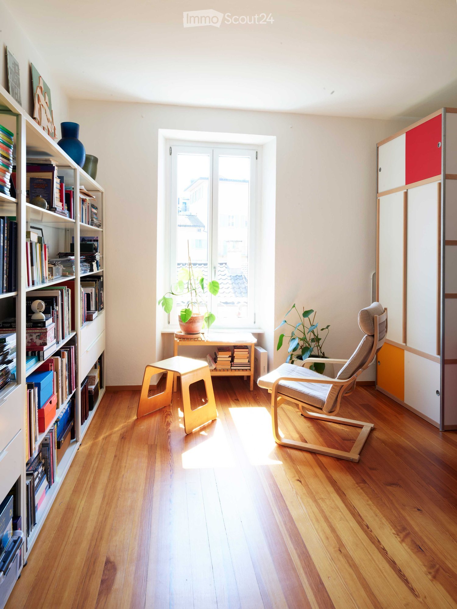 Bright, minimalist room with wooden flooring, a large window, a bookshelf filled with books and decorative items, a small table with a plant, a modern chair, and a small stool.