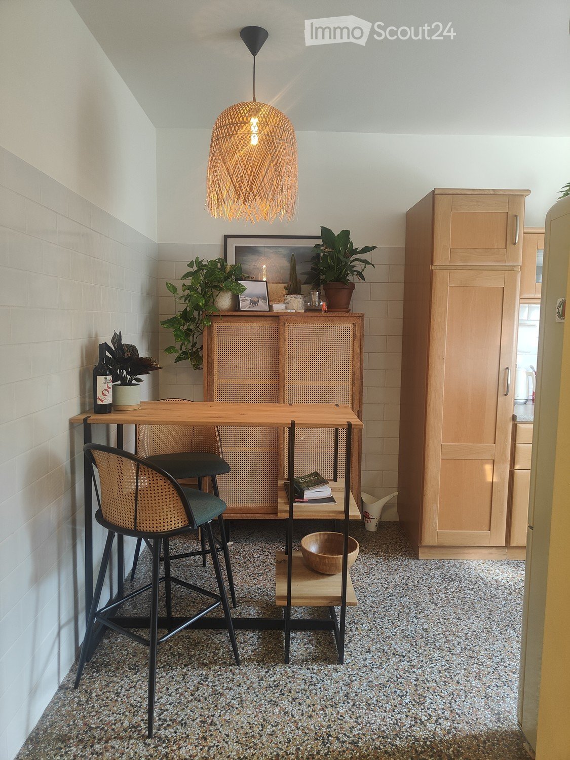A modern, compact dining area with a wooden table, two wicker chairs, a pendant light, and a cabinet with a woven front. The space includes plants, a bottle, and a bowl on the floor.