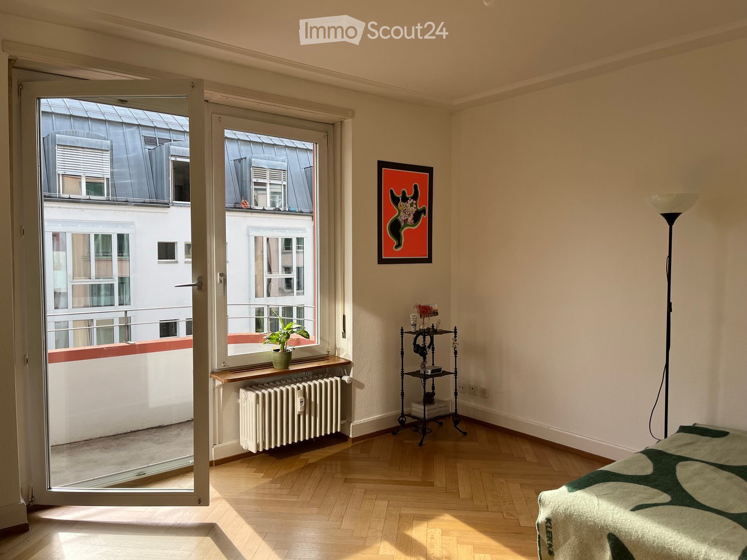 Bright room with wooden flooring, large window opening to a balcony, radiator, small table with plant, picture frame on wall, floor lamp, and bed with green and white patterned bedspread.