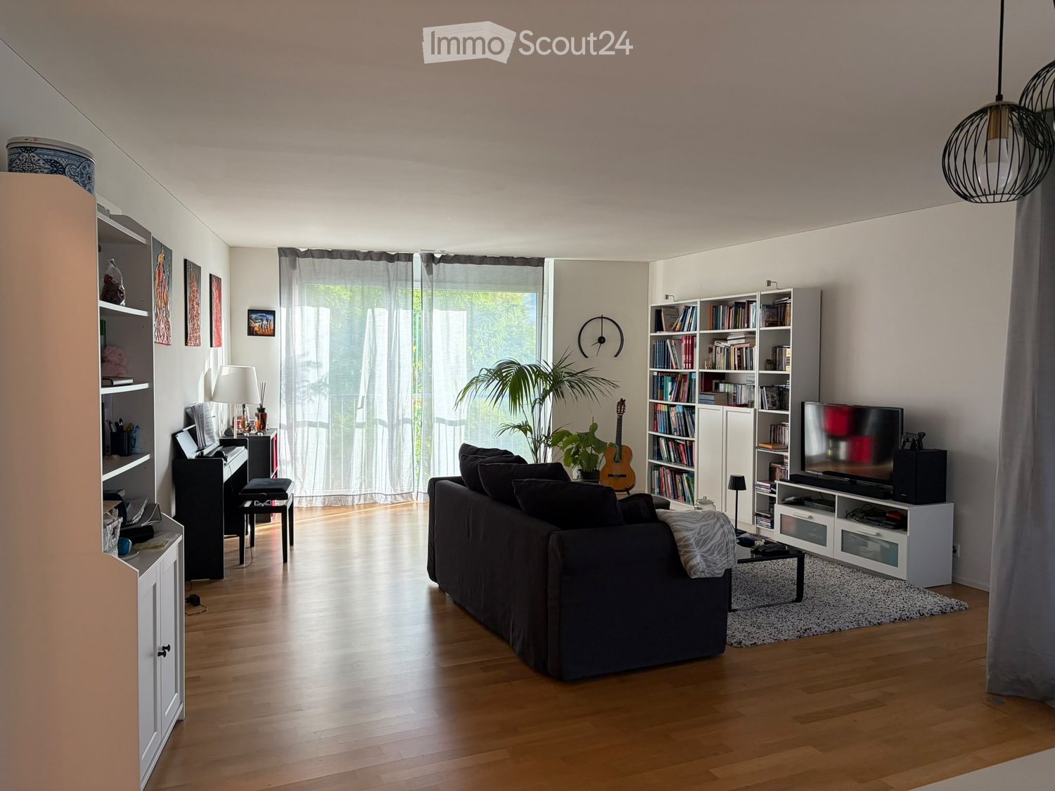 Modern living room with wooden flooring, large windows, a dark-colored sofa, coffee table, bookshelf, piano, and a television stand.