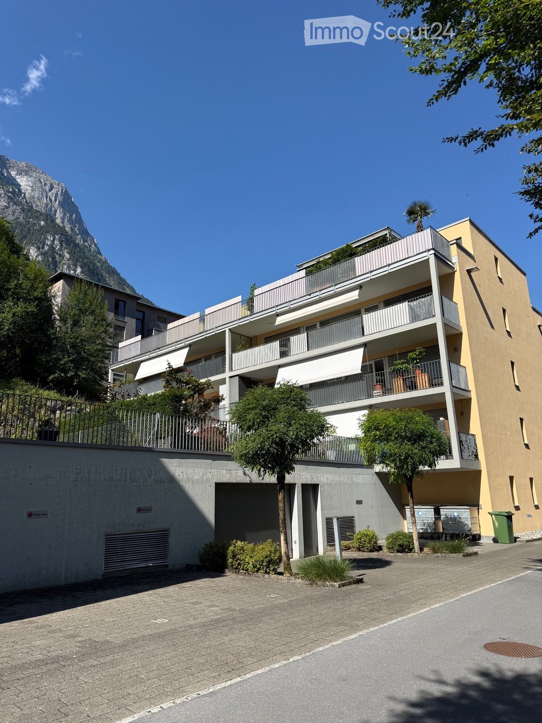 Modern multi-story residential building with balconies, surrounded by trees and a clear view of a mountain in the background.