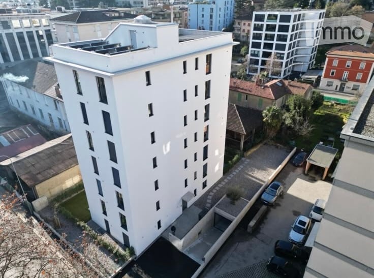 A modern, multi-story residential building with a flat roof and numerous windows, surrounded by other buildings and a parking area.