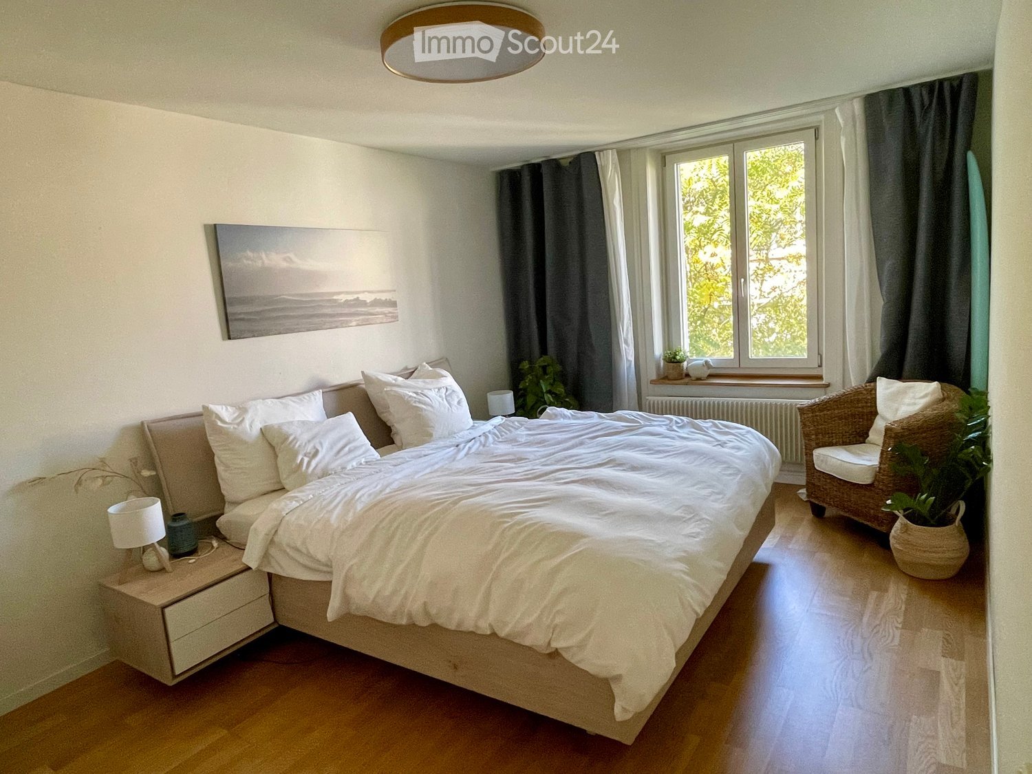 Cozy bedroom with a large bed, nightstand with lamp, wicker chair, potted plant, and a scenic picture on the wall.