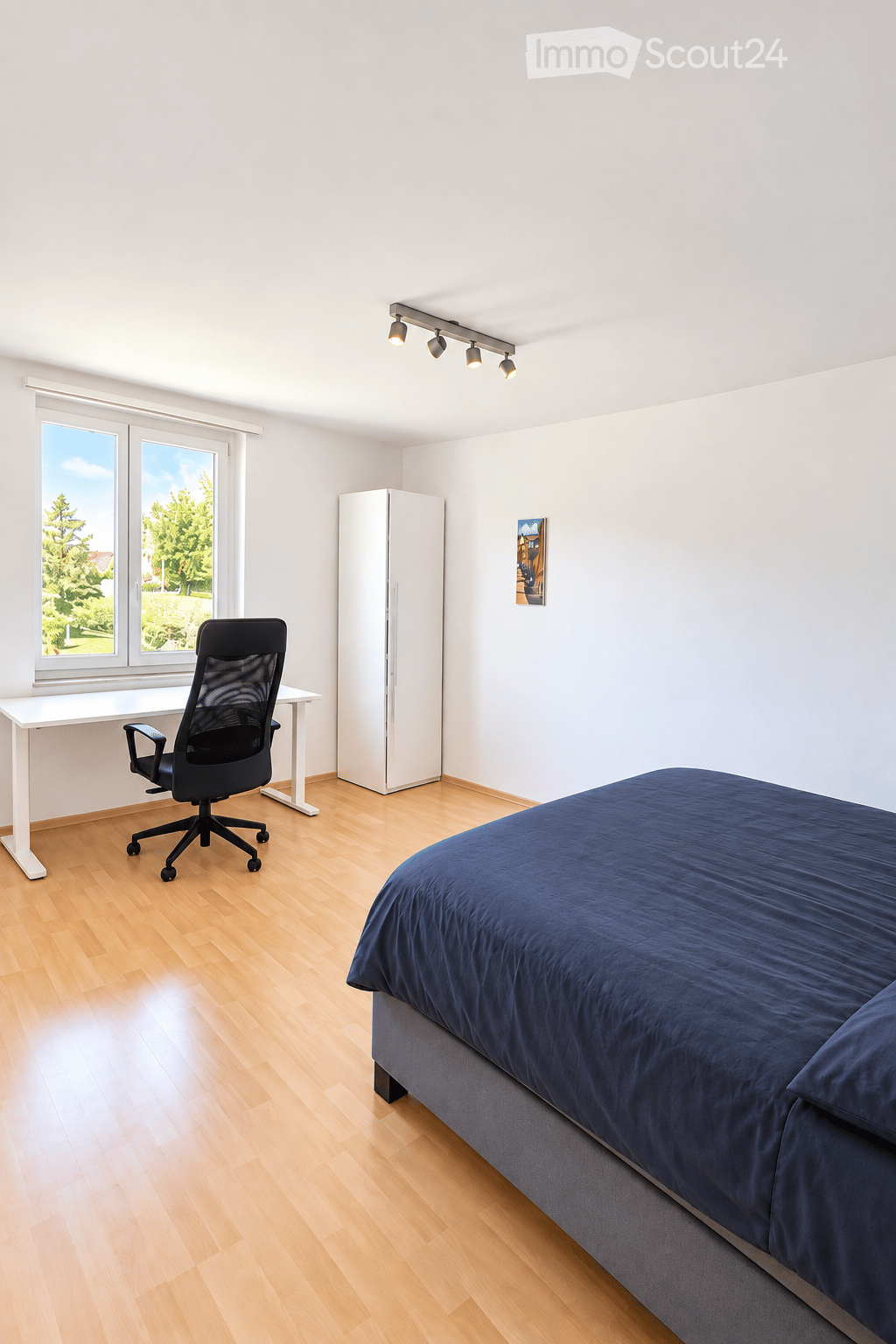 Bright bedroom with wooden flooring, large window, bed with dark blue bedding, desk with chair, and a white wardrobe.