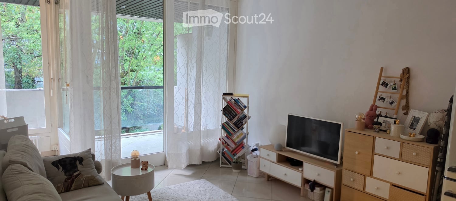 Bright living room with large windows and balcony access, featuring a sofa, coffee table, bookcase, TV, and decorative items.