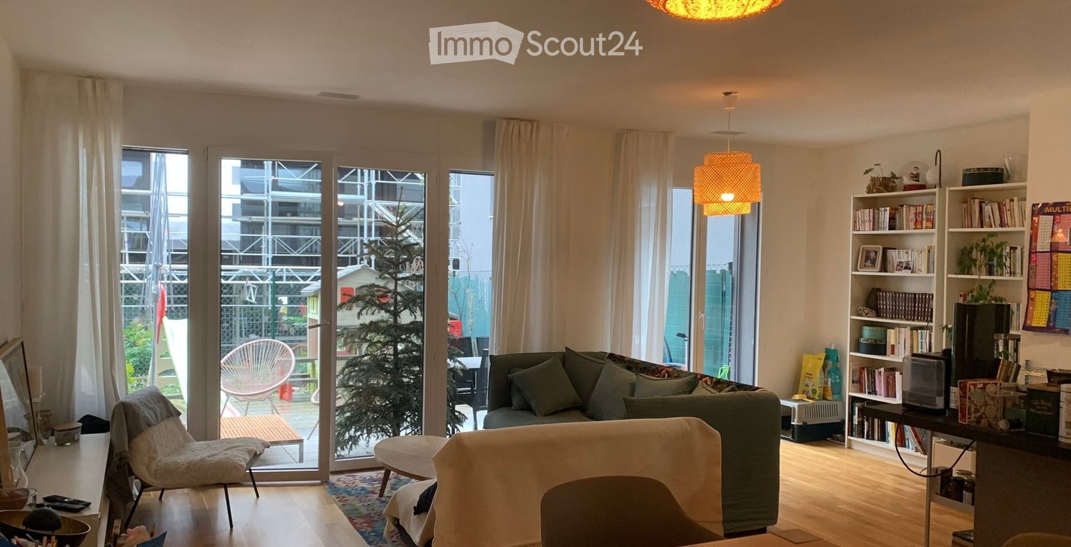 Bright living room with large windows and sliding glass doors leading to a balcony. Features a green sofa, a white chair, a small table, a bookshelf, and a hanging lamp.