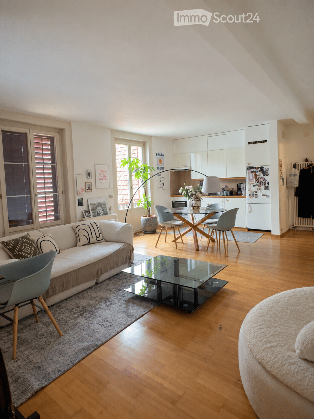 Modern living room with a white sofa, glass coffee table, dining table with chairs, kitchen area with white cabinets, refrigerator, and a potted plant. The room features wooden flooring and large windows with blinds.