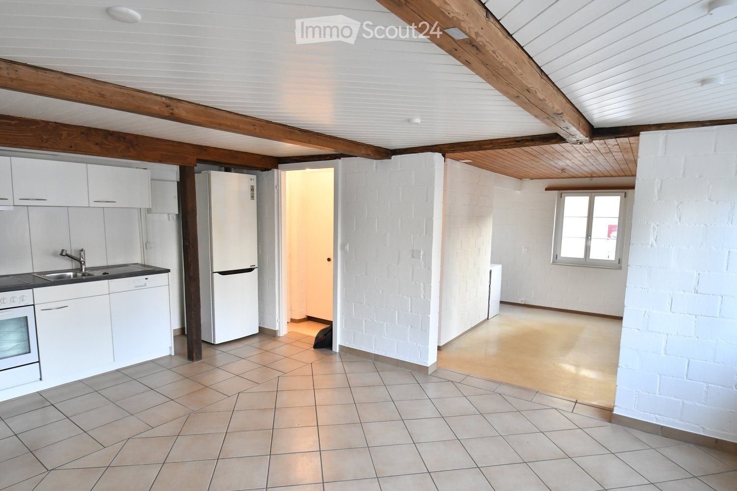 Bright, open-plan room with exposed wooden beams, white brick walls, and tiled flooring. Includes a kitchen area with white cabinets, a refrigerator, sink, and oven, connected to a larger living space with large windows.