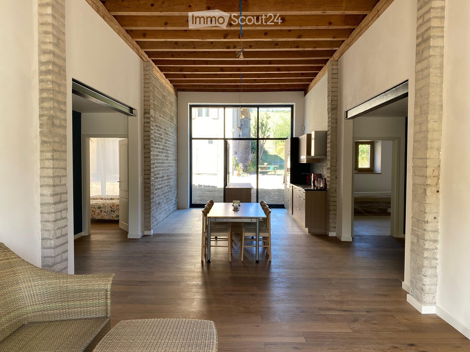 Open-plan living and dining area with wooden flooring, exposed wooden ceiling beams, brick walls, and large glass doors leading to an outdoor space. Visible furniture includes a sofa, dining table with chairs, and kitchen cabinets.