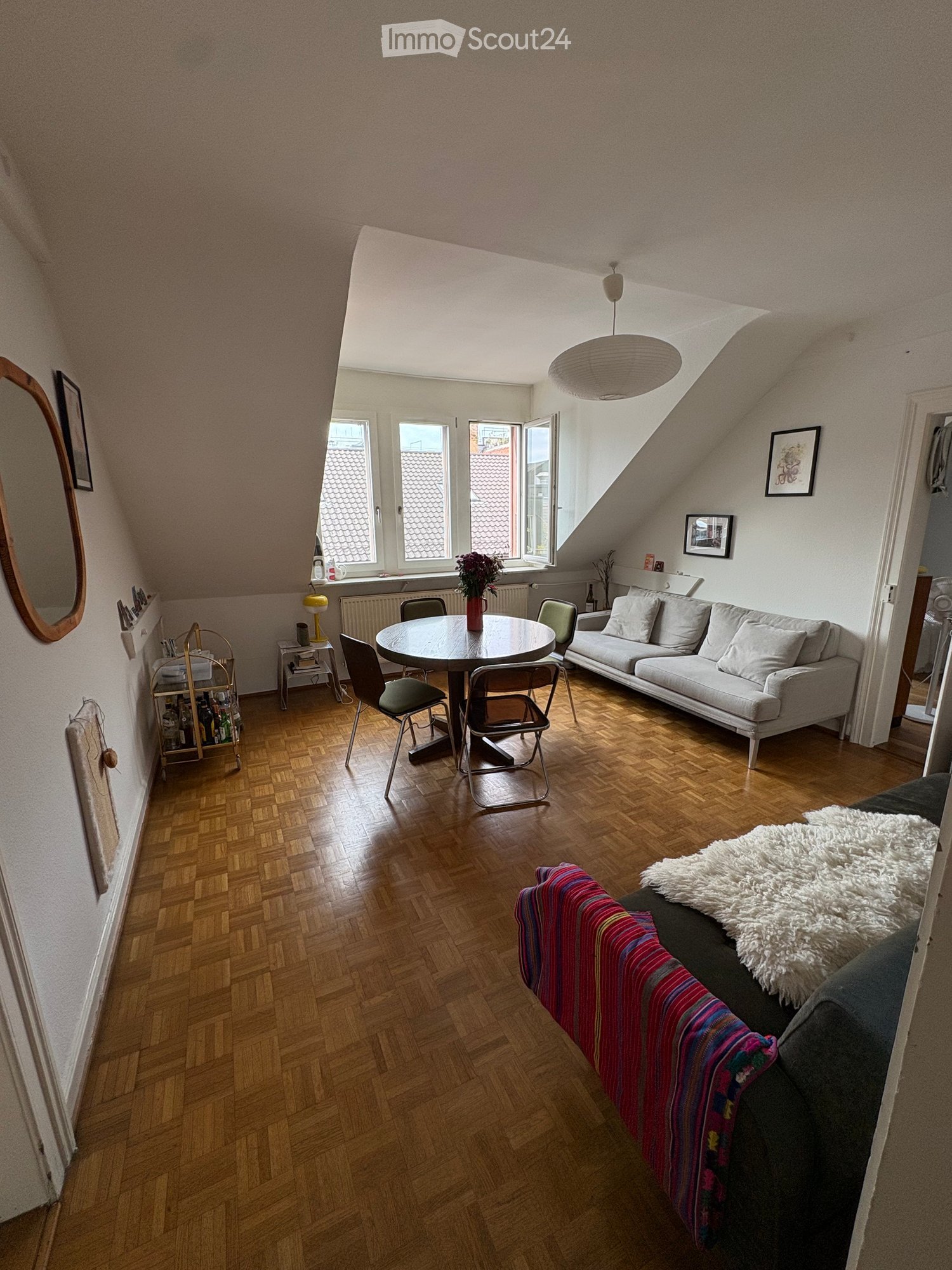 Cozy living room with wooden flooring, large windows, a grey sofa, a round dining table with chairs, a bar cart, and decorative items including a mirror and picture frames.
