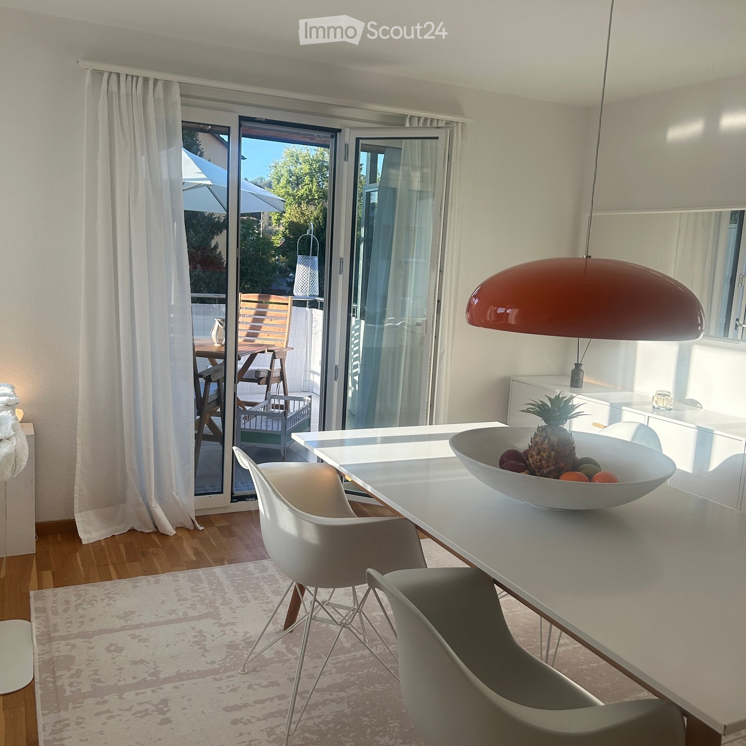 Modern kitchen-dining area with white furniture, a large window with a view, and a bowl of fruit on the table.