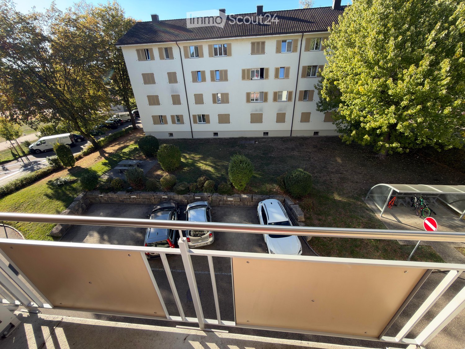A multi-story residential building with numerous windows and shutters, surrounded by greenery and a parking area with several cars.