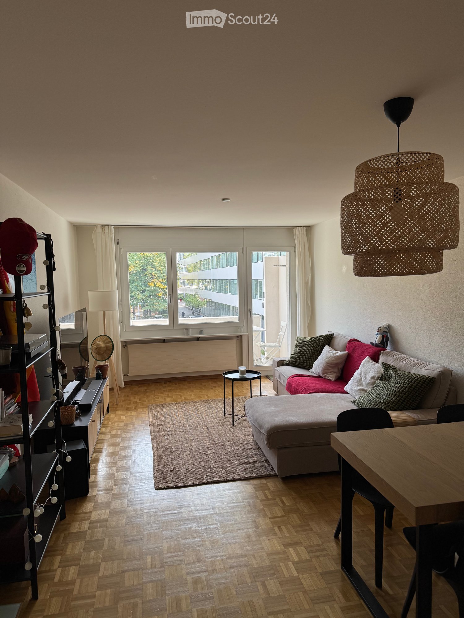 Modern living room with wooden flooring, large windows, a sofa with colorful cushions, a small round table, and a shelf with decorative items.